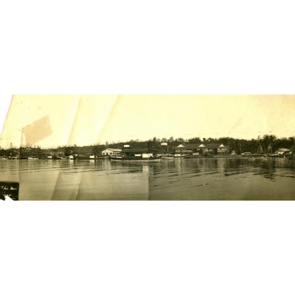 A panoramic photo of a buildings at the edge of a waterway. There are boats on the water in the foreground and oil rigs along the hills in the background.