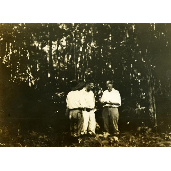 A photo of three men wearing white, standing in the jungle. The man on the left is facing away and is wearing a hat.