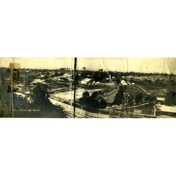 A panoramic photo of a town with buildings along a coast line. Water can be seen in the background. There are oil derricks on the left and right.