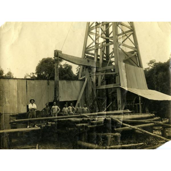 Une photo de huit hommes debout derrière une pile de tubage et devant une installation de forage pétrolier recouverte de tôle. 