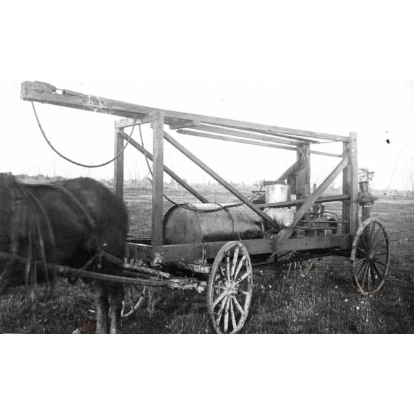 Un cheval tirant une charrette à quatre roues. Il y a une poutre en bois qui dépasse au-dessus d'un cadre en bois ouvert et sur lequel repose un tonneau en métal.  