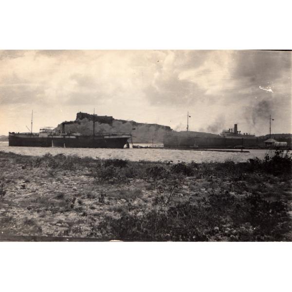 A photo of two ships tied to a dock on a pier. There is a hill in the background and gravel and plants in the foreground. 