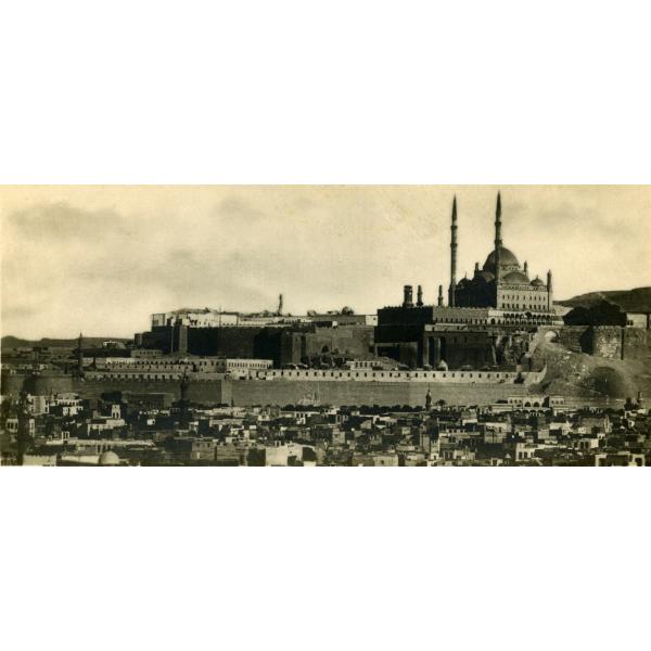 A photo of a large building surrounded by a tall stone wall. There is a building with a dome sitting up on a hill behind it and smaller buildings in front of the wall.