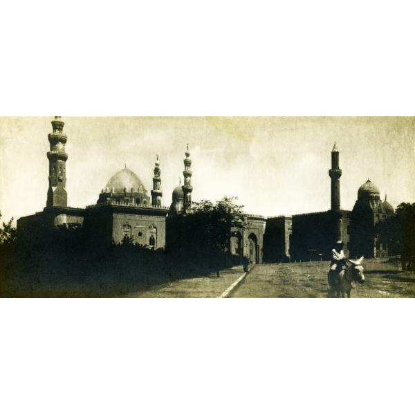 A photo of a building with a domed roof and four towers. There is a person riding a donkey in the road leading up to the building entrance.