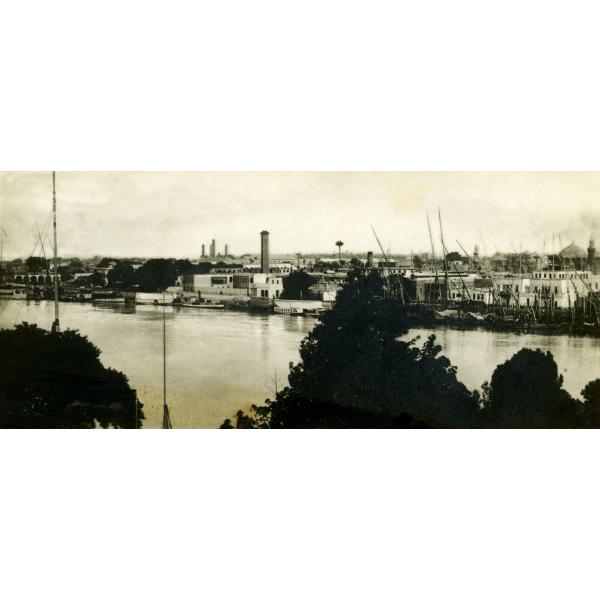 Photo d'un bâtiment à l'autre bout d'une rivière, avec le sommet d'un arbre au premier plan. Des bateaux avec de grands mâts amarrés le long du rivage.