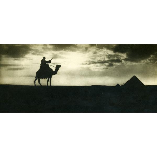 A photo of a man riding a camel and holding a cane. There are two pyramids to the right. The sky behind is full of clouds and the sun in shining down, casting the man and pyramids in shadow.