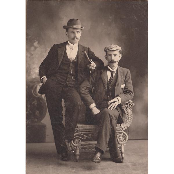 Charles Wallen leaning on a chair that Alf Burns is sitting in. They are wearing dark suits and hats, and are holding pipes. The background is a dark wall. 