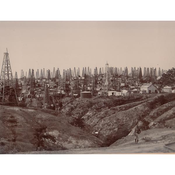 A photo of ten oil derricks close together on a hill. Some have cylindrical, steel storage tanks. There is a steam boiler, with tall stacks, in the centre. 