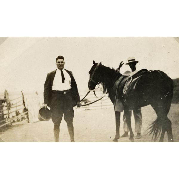 George Rawlings in a dark suit and riding boots. He is holding a pith helmet in one hand and the reins of a horse in the other. A man is standing behind the horse.  