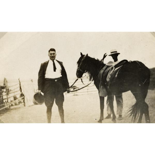 George Rawlings dans un complet foncé et des bottes d’équitation. Il tient un casque colonial dans une main et les reines du cheval dans l’autre. Un homme se tient derrière le cheval.  