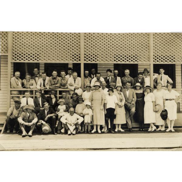 A group of International Drillers and their families outside of a building with latticework on the porch. The men are wearing suits and the women are wearing dresses. 