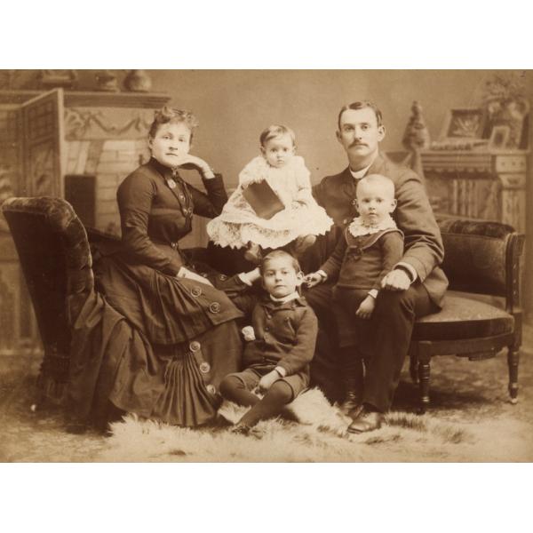 A woman and man sitting with three children between them. They are wearing dark clothing except for the youngest child in the middle who is wearing white. There is a mantle behind them and a fur rug at their feet. 