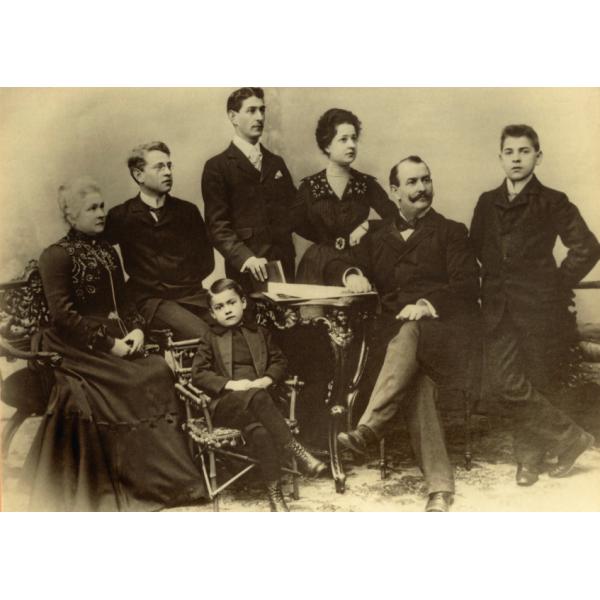 A posed photo of eight people, looking off to the right. They are wearing dark clothing. The two women (far left and third from right) have embroidery on the tops of their dresses. 