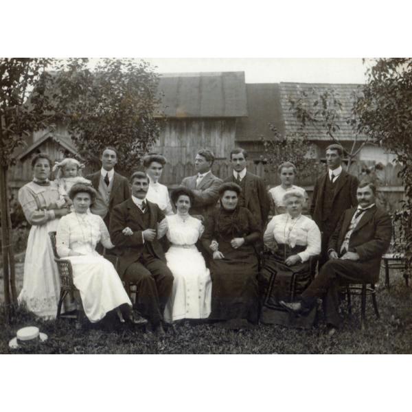 A photo of eight people standing and six people sitting in chairs in front of a building and trees. 