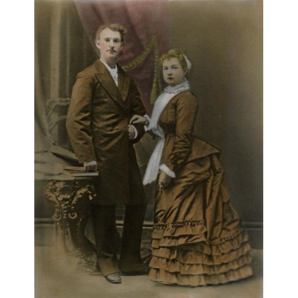 A man and a woman standing beside a table and in front of a purple curtain. He is wearing a long brown jacket and pants. She is wearing a long brown dress with a white apron over top and is holding is arm with her hand.