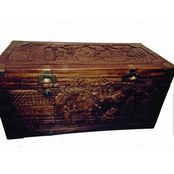 The front of a carved cedar chest. There are scenes carved into the lid and side showing people, trees, and buildings. There are golden corners and openers on the box. 