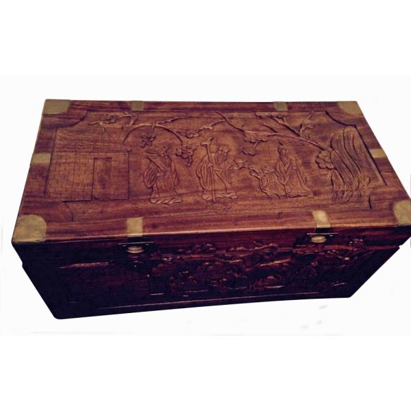 The top of a carved cedar chest. It depicts three men, one with a staff who is handing something to a seated man while the other looks on. There is a building on the left. 