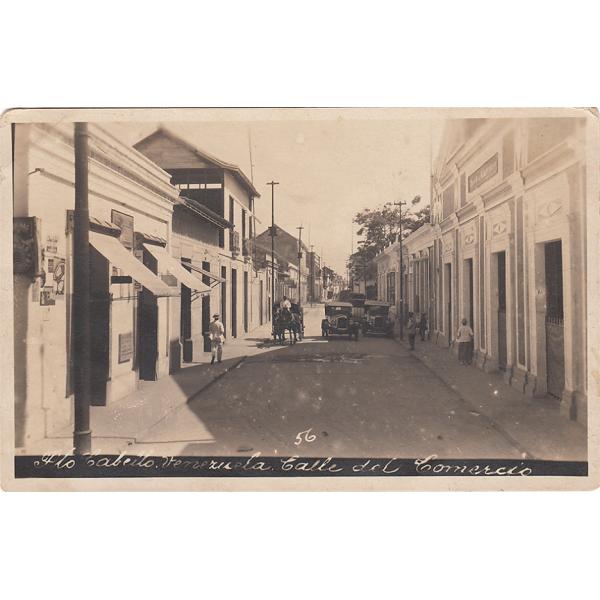 The front of a postcard showing a street with cars and a team of horses in the road.