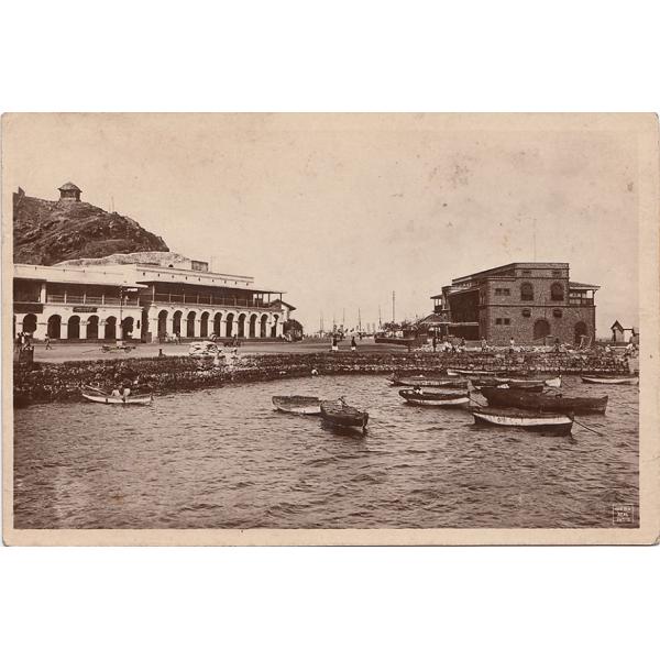 The front of a postcard showing small fishing boats along the edge of the water and two large buildings behind them on land. 