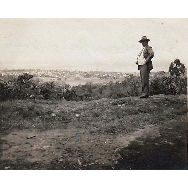 Humphrey Tracy debout sur une colline, regardant Port d'Espagne. Il porte un costume et un chapeau. Il y a des arbres et de l’eau en arrière-plan.