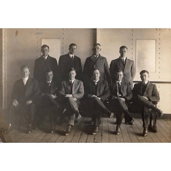 A photo of four International Drillers standing and six sitting in chairs on a boat deck. They are wearing dark suits and ties. They are in front of a wall that has five columns of rivets in it. 