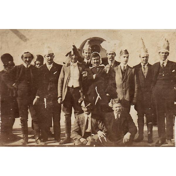 A group of men wearing dark suits. Eleven are standing and two are sitting. The men are wearing paper hats. 