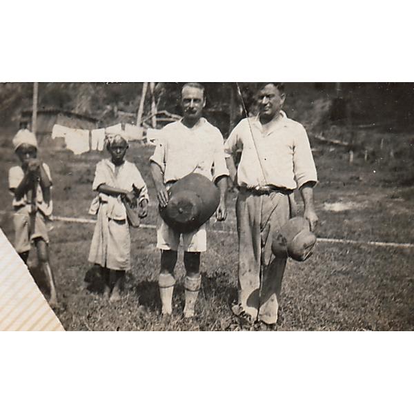 Two native boys standing beside Tracy and Hicks, who are wearing light-coloured clothing and are holding hats. 