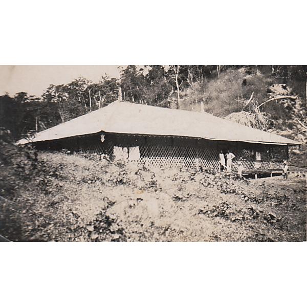 A bungalow set into a hill with a light roof and large latticework surrounding it. Three people stand on the stairs and another stands a few feet away.  