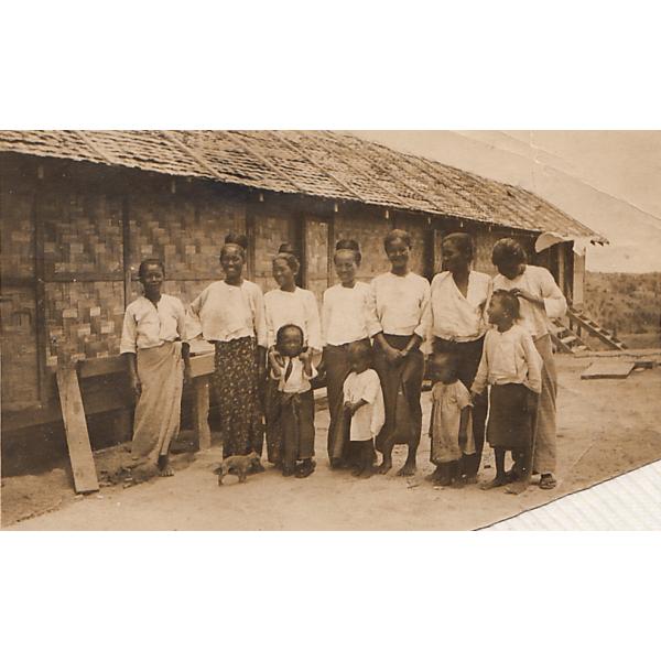 Une photo de sept femmes et quatre enfants se tenant devant un édifice en chaume. Les femmes portent des jupes et des chemisiers blancs. 