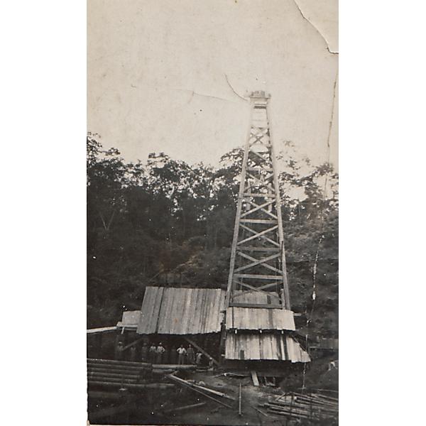 A photo of a covered oil rig. There is a group of men in front of it. There is casing in the foreground and trees in the background. 