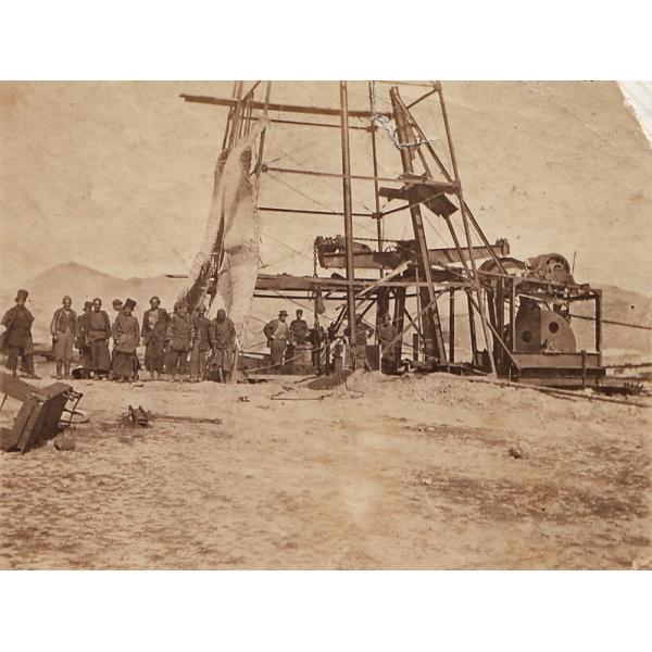 A photo of a group of men wearing work clothes stand at the base of a metal oil rig. There is a piece of cloth covering the left side of the rig. The ground is bare and there are hills in the background. 