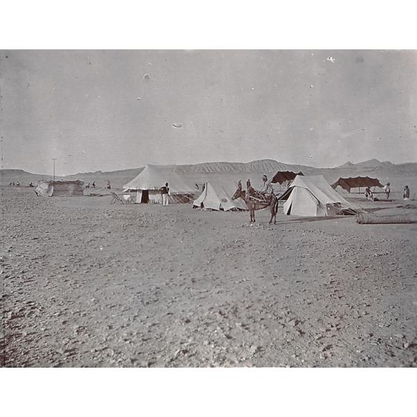 A photo of three white tents in the desert with two black tents behind. There are several people around the tents and one riding a donkey in the foreground. 