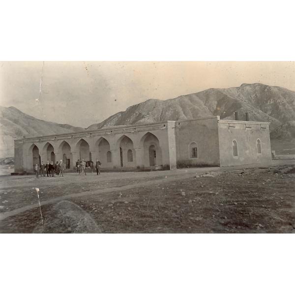 Une photo d’une maison de plain-pied avec un toit plat et sept arches à l’avant. Un groupe de personnes et un cheval se tiennent devant celle-ci, avec des collines à l’arrière. 