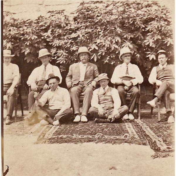 A photo of three patterned rugs on the ground. Five International Drillers are sitting in chairs and two are sitting on the ground. There are bushes behind them. 