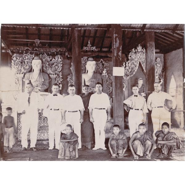 Six International Drillers wearing white inside a temple with five young boys and a monk. There are three pillars running from the floor to the ceiling and three statues in the background. 