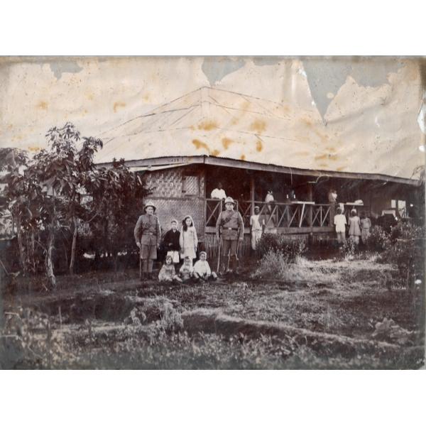 A photo of a thatched bungalow. There is a group of people outside the house, and two of the men are in uniform. There are trees on either side of the photo. 