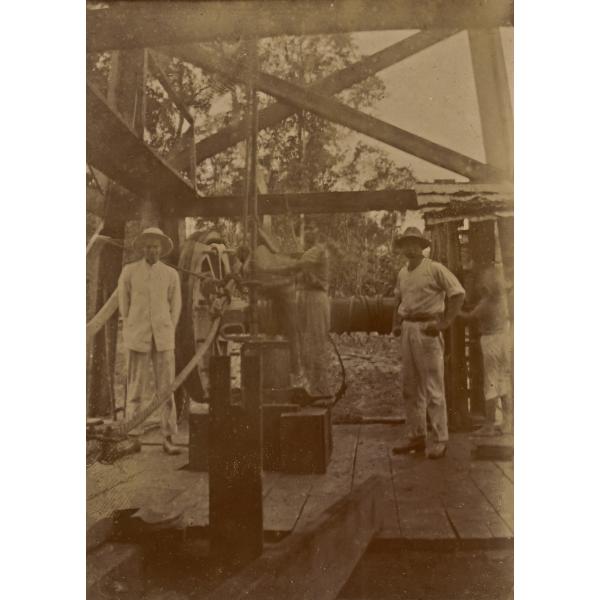 Bloss Sutherland and two men standing under an oil rig. There is a large wheel connected to a belt behind them and trees in the background. 