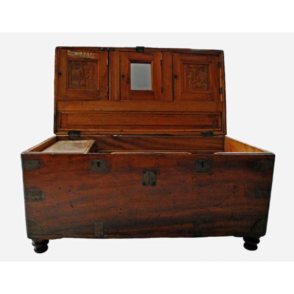 A smooth wooden chest with a reddish hue with the lid open. There are pieces of brass on the corners and there are circular wooden feet. The lock is made of brass. The lid has two side compartments with a carved flower design. The centre compartment has a mirror on it. The body of the chest has a raised compartment on the left and the right side is hollow.