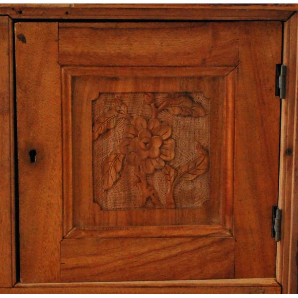 A compartment door on a smooth wooden chest with a reddish hue. There is a flower carved into it and there are black hinges on the right. There is a small keyhole on the left,