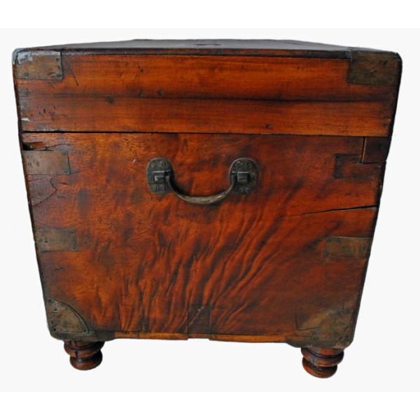 The side of a smooth wooden chest with a reddish hue. There are pieces of brass on the corners and the handle is also made of brass. There are circular feet on the bottom of the chest.