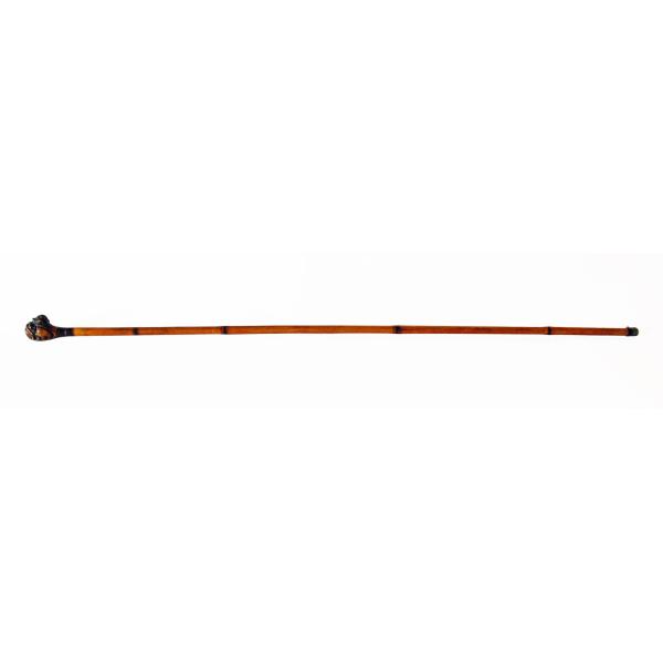 A bamboo walking cane with an orange shaft. There is a carved monkey on the handle and a metal stopper on the bottom.