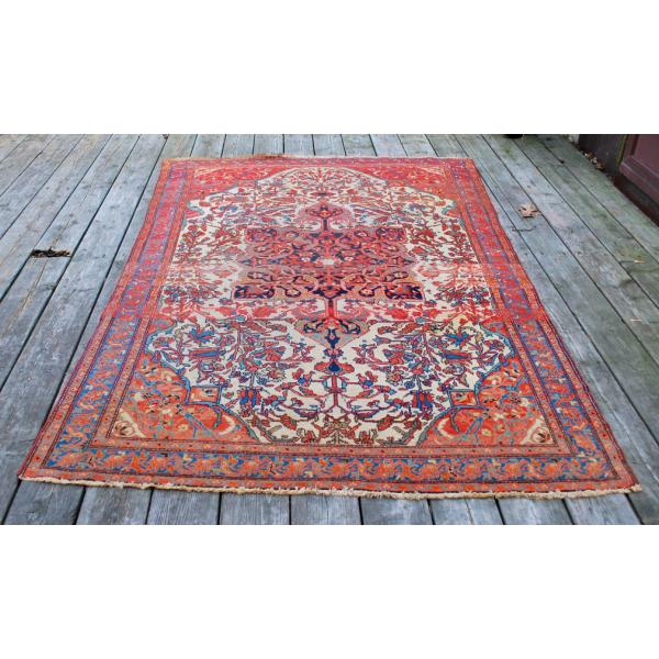 A rectangular red rug with a blue, white, orange, green, and yellow motif in the centre. It is sitting on a wooden deck. 