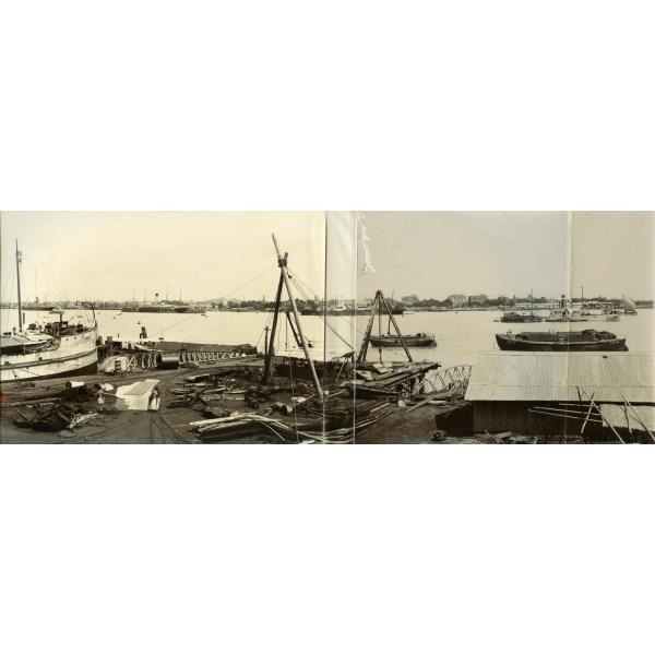 A photo of boats tied up to a dock. There are building materials and metal tracks on the dock. More boats float out in the water behind and there are buildings along the far bank of the water.