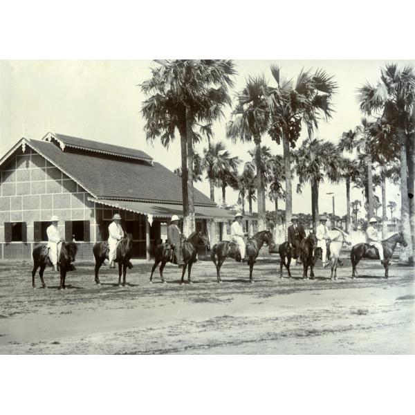 A photo of seven International Drillers lined up on horses facing right. They are wearing pith helmets. There is a buildings with large tiles in the background, and a group of palm trees.