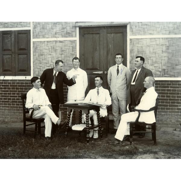 Four men standing behind three men seated in chairs around a small table. There is a building with brick on the bottom part of the walls, with wood details on the upper parts. The ground is covered in grass.