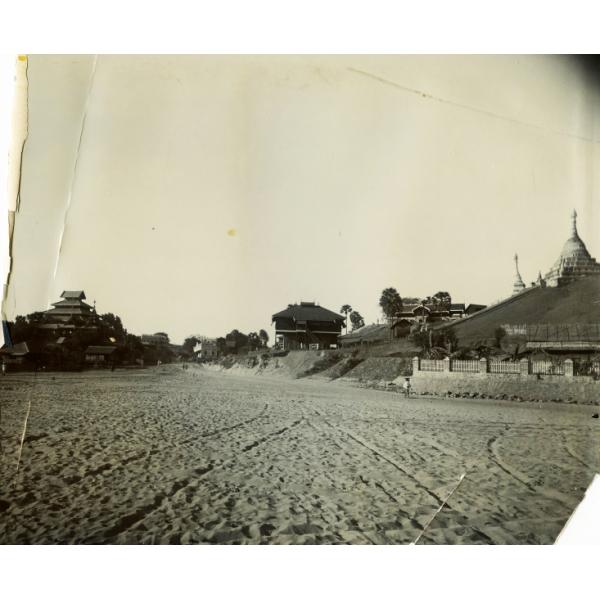 A photo of a group of buildings sitting on an embankment on either side of a sandy road. The buildings on the left and right have tiered roofs.