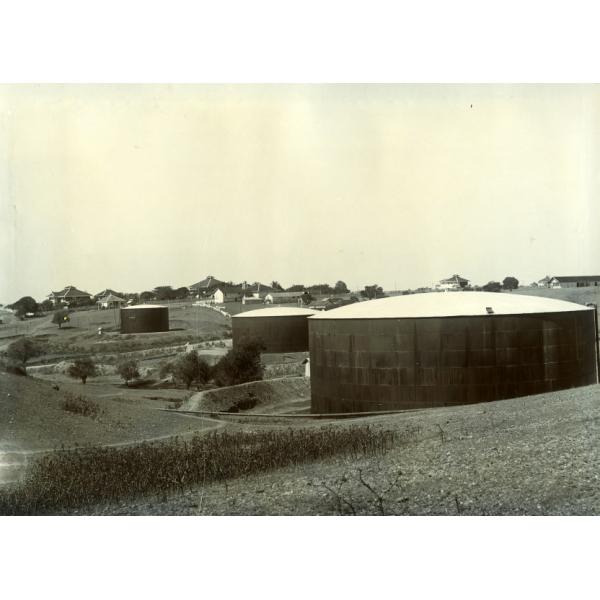 A photo of three oil storage tanks in front of a number of buildings. The sides of the tanks are made from black panels and the roofs are white.