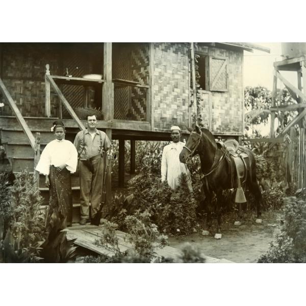 Harry Phillips se tient entre une femme birmane et un homme devant les marches d'une maison sur pilotis. Il tient un fusil et porte des bretelles. La femme porte une longue jupe. L'homme tient un cheval foncé. 