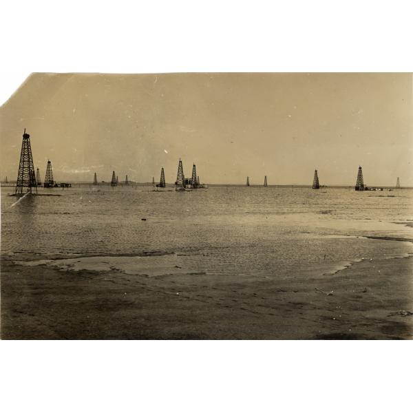 A photo of water washing over the bottoms of oil derricks that are built on a sandy beach. 