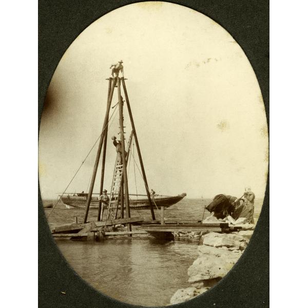 Une photo de trois hommes sur une tour en bois avec quatre poteaux et des cordes descendant jusqu'à un quai. Un bateau au mât abaissé est amarré dans l'eau derrière eux. Un quatrième homme se trouve à droite à côté d'une pile d'objets enveloppés dans une matière foncée.  